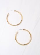 Pair of large gold tubular hoop earrings with post backs on marble surface